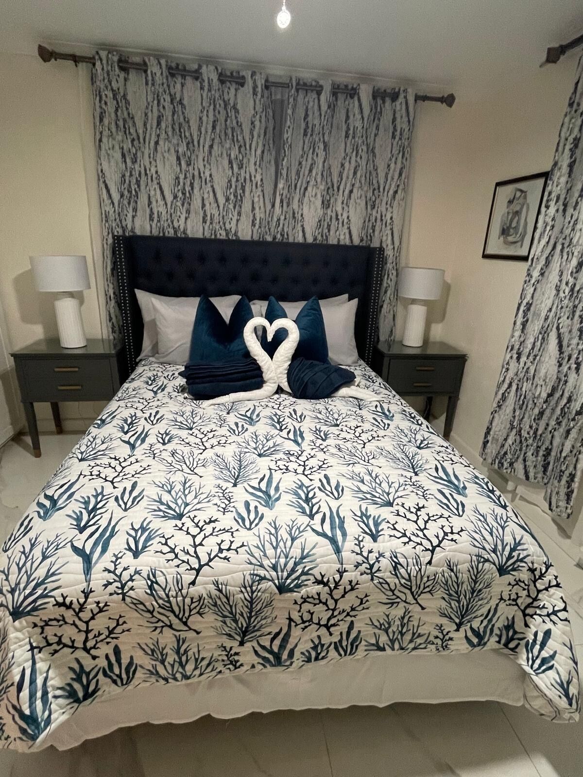 A bed with a blue and white coral pattern bedspread, two heart-shaped pillows, a dark headboard, and matching nightstands with lamps.