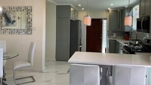 A modern kitchen with a central island, pendant lighting, and a dining area with a glass table and chairs. - K&L Villa Apartment (Christ Church)