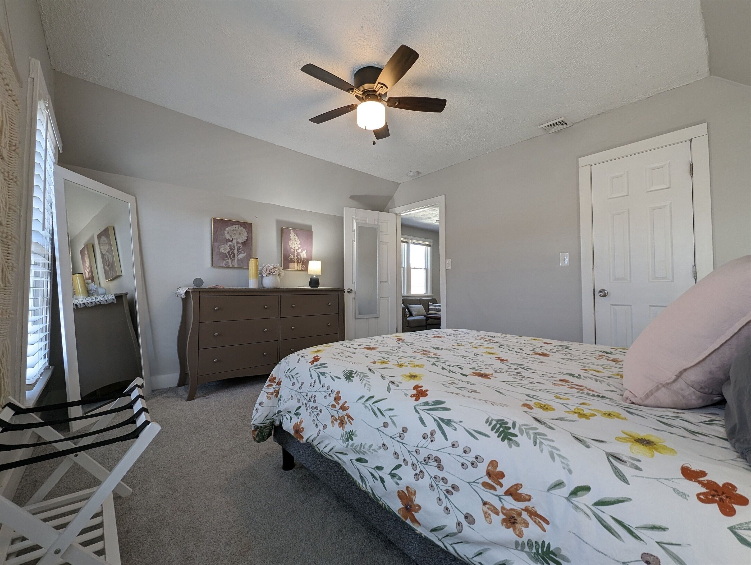 Une chambre à coucher comprenant un lit, une commode, un ventilateur de plafond et une porte donnant sur une autre pièce.