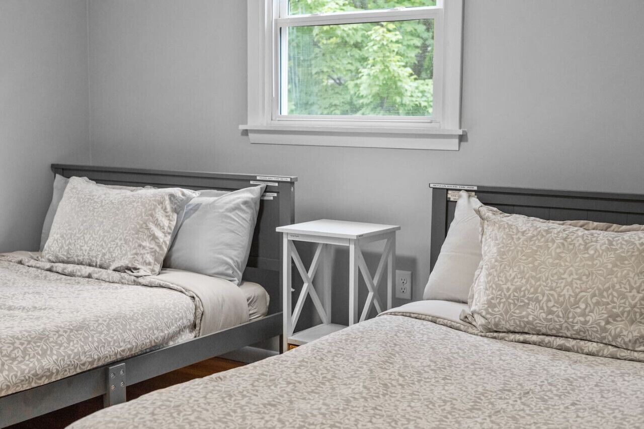 Un dormitorio con dos camas, una mesita de noche pequeña y blanca, y una ventana con vistas a árboles.