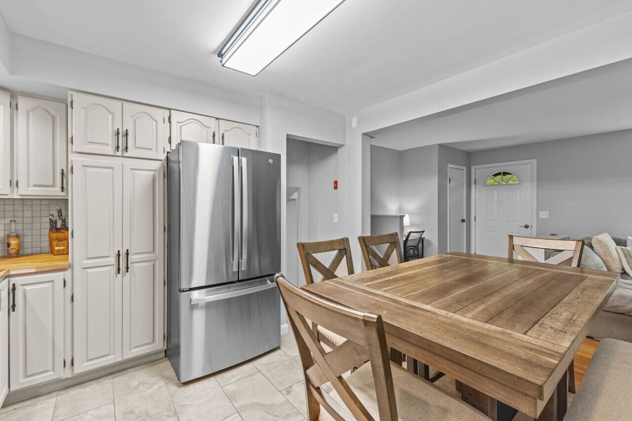 A modern kitchen with white cabinetry, a stainless steel refrigerator, and a wooden dining table with chairs.