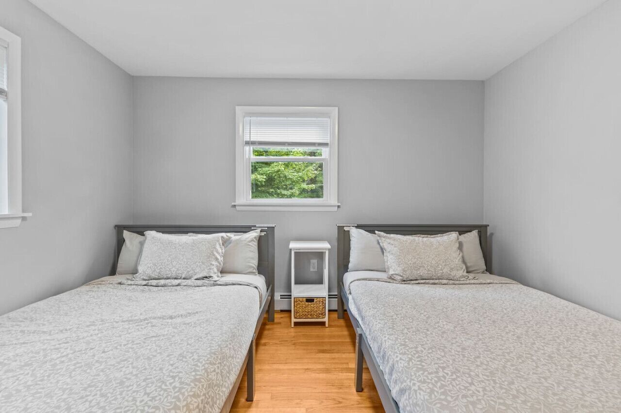 Une chambre avec deux lits simples, une fenêtre avec des stores et une petite table de chevet avec un panier.