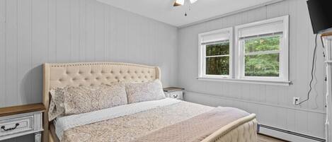 A bedroom with a tufted headboard, a bed with patterned bedding, a nightstand, a bedside table, a ceiling fan, and a window with a view of greenery.