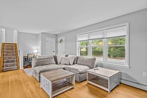 A modern living room with a sectional sofa, two side tables, a staircase with a patterned rug, and a large window with blinds. - Charming 5B Home in Neighborhood (South Burlington)