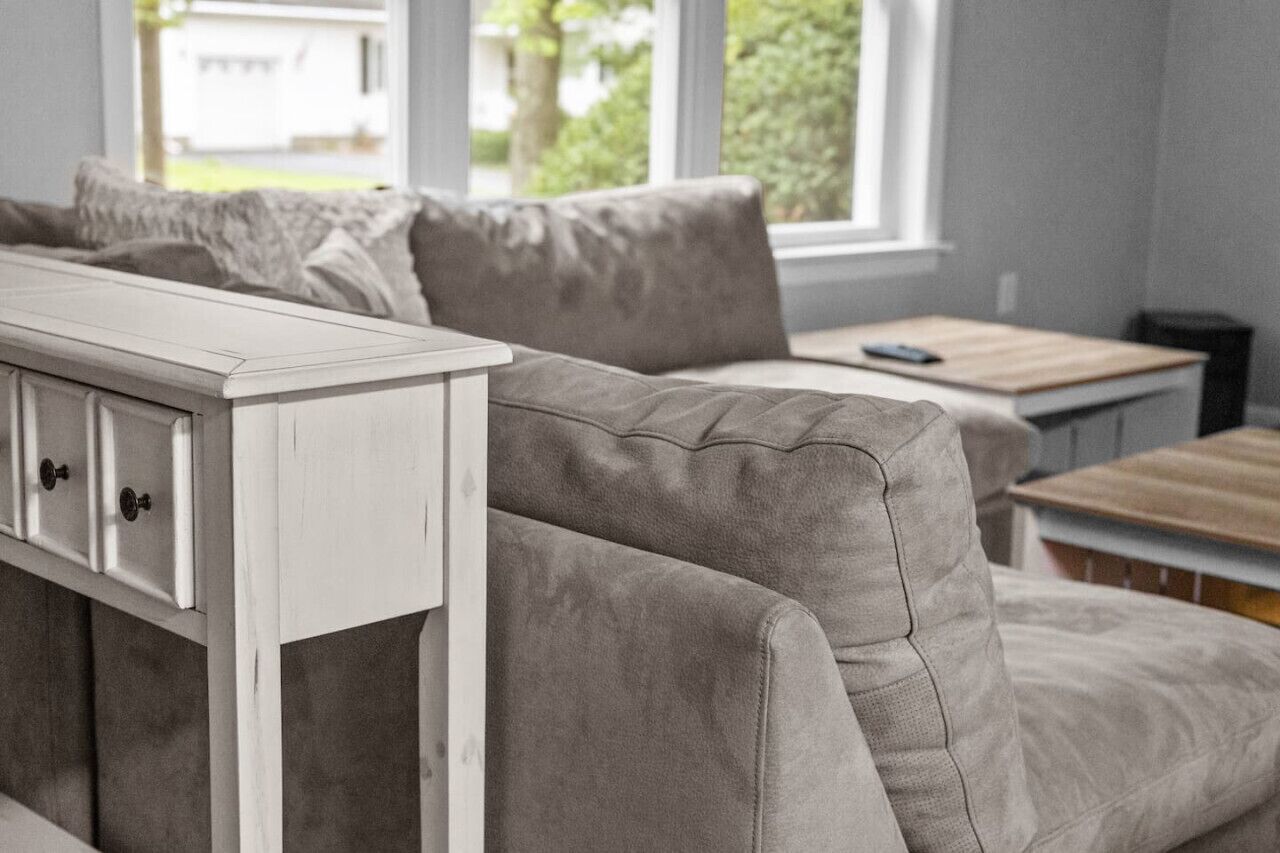A neutral-toned living room with a sofa, a white side table, and a window with a view of greenery.