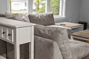 A neutral-toned living room with a sofa, a white side table, and a window with a view of greenery. - Charming 5B Home in Neighborhood (South Burlington)
