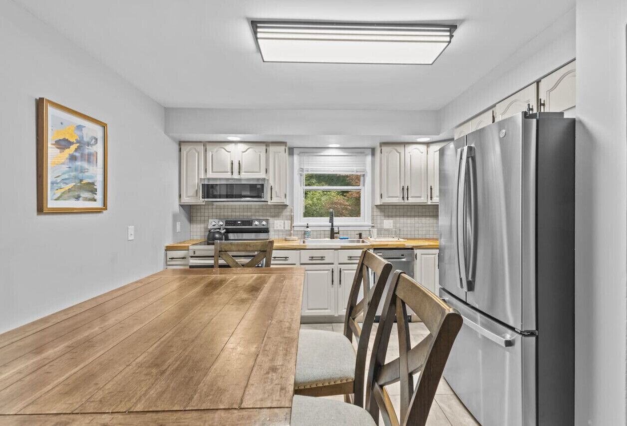 A modern kitchen with white cabinets, stainless steel appliances, a wooden dining table, and a window with blinds.