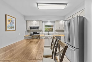 A modern kitchen with white cabinets, stainless steel appliances, a wooden dining table, and a window with blinds. - Charming 5B Home in Neighborhood (South Burlington)