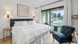 Quarto de hotel com uma cama grande, duas luminárias laterais, um ventilador de teto, uma poltrona azul escura e vista para o exterior através de uma porta de vidro deslizante.