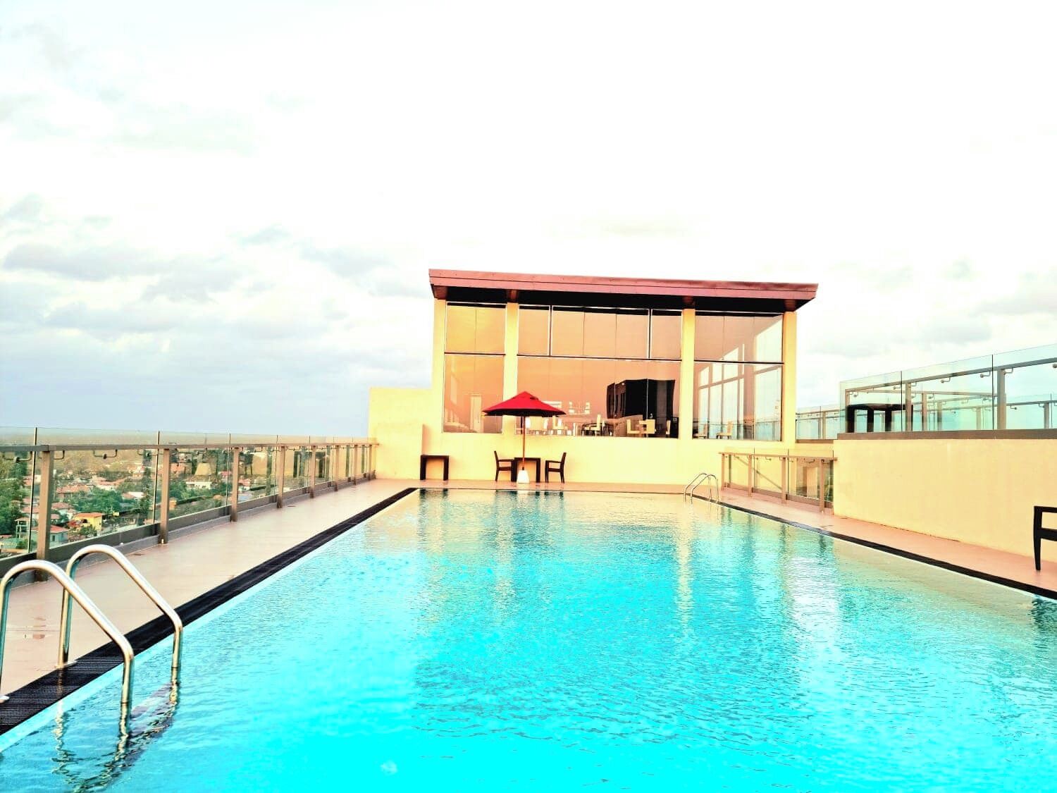 A rooftop infinity pool with a glass balustrade, a red umbrella, and a modern building with large windows.