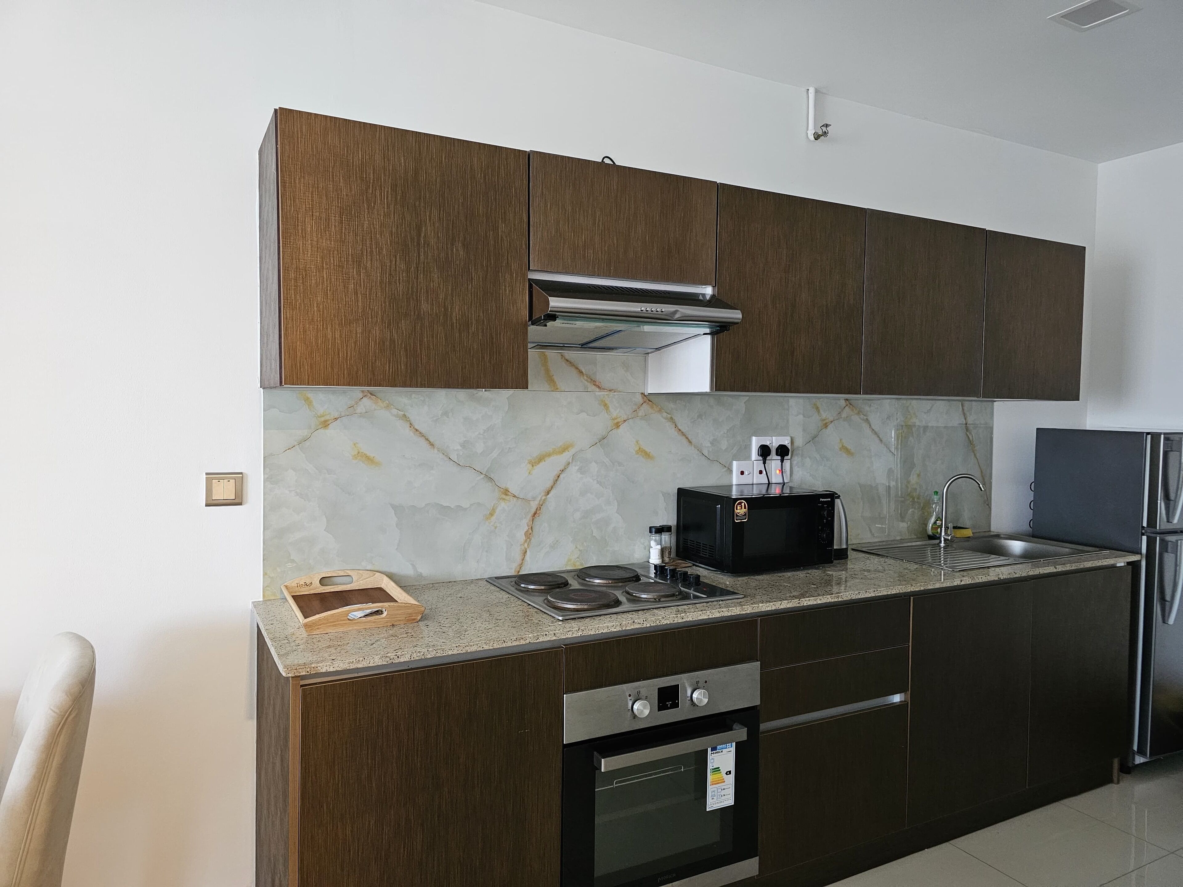 A modern kitchen with dark wooden cabinets, a stainless steel refrigerator, a built-in oven, and a marble backsplash.