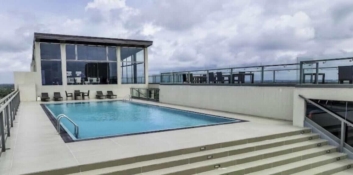 Un área de piscina en la azotea con un salón envuelto en cristal, una piscina y una escalera que lleva a una terraza.