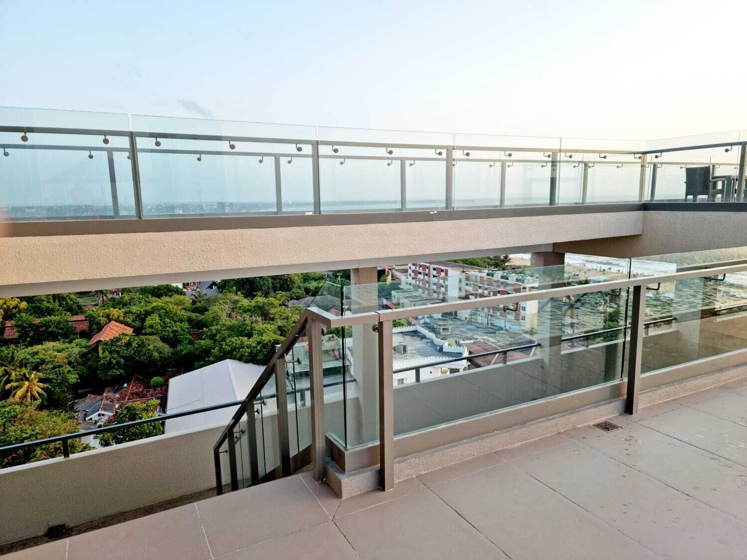 En udendørs terrasse i flere niveauer med glasgelændere, der giver udsigt over et boligområde og en bro i det fjerne.