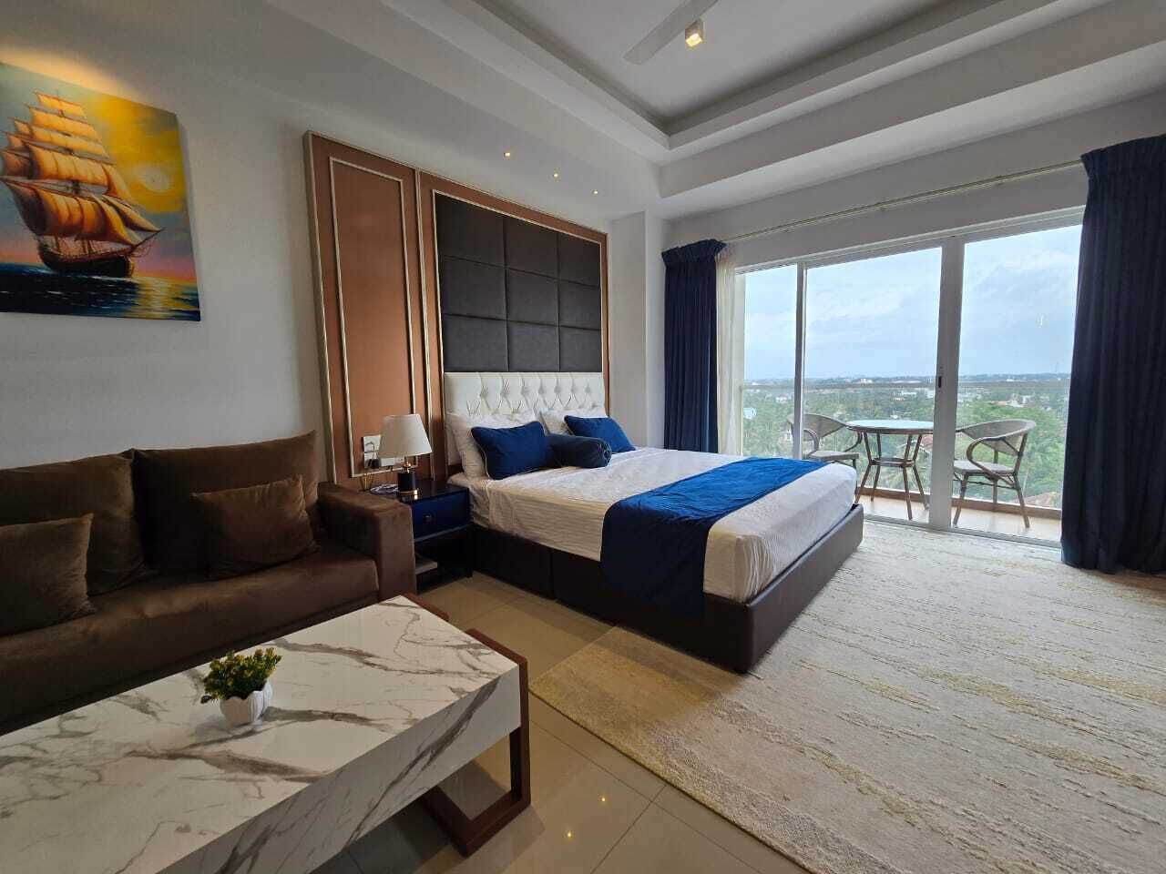 A modern hotel room with a large bed, a sofa, a small table, and a balcony with a view.