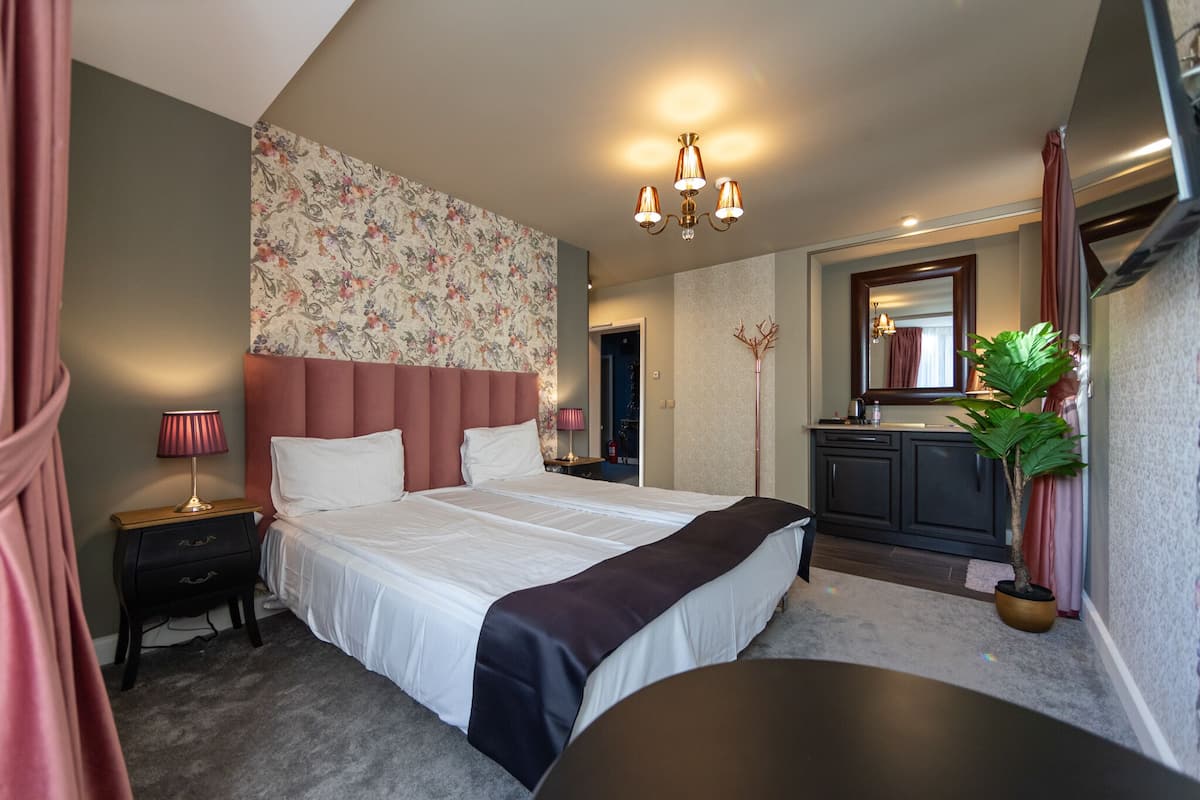 Ein Schlafzimmer mit einem großen Bett, Nachttischen, einem Schreibtisch, einem Stuhl, einer Zimmerpflanze, einem Spiegel und einem Badezimmer mit Waschbecken und Schrank.