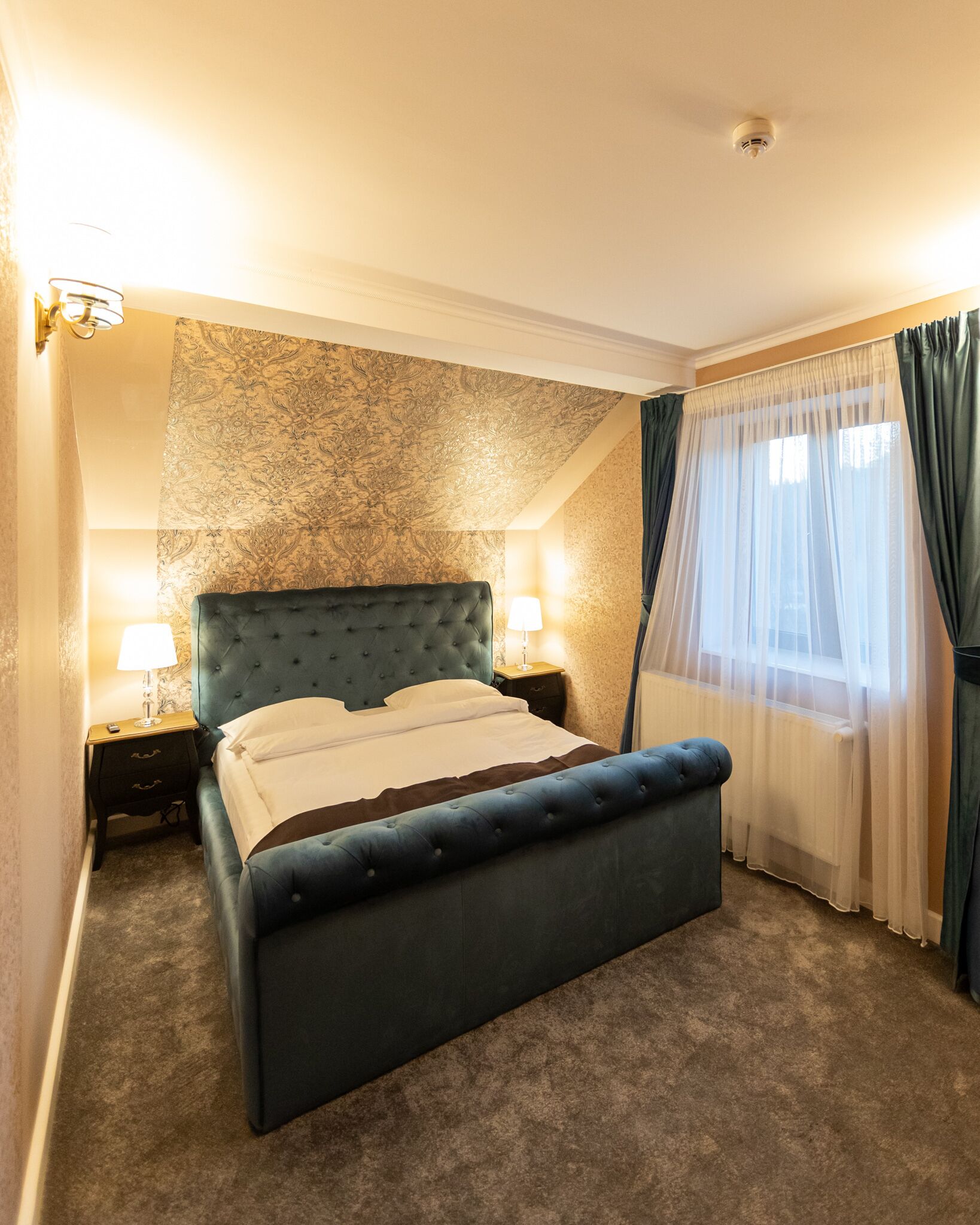 Ein Schlafzimmer mit einem großen Bett, zwei Nachttischen mit Lampen, einem Fenster mit Vorhängen und einer Wand mit Muster.