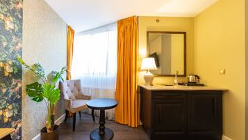 Une chambre d’hôtel avec un papier peint à motifs floraux, une plante en pot, une chaise, une petite table ronde, un lavabo avec un miroir et une fenêtre avec des rideaux.