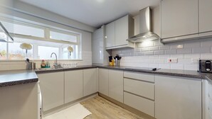 A modern kitchen with white cabinets, a stainless steel range hood, and a window with flowers. - Lovely 3 Bedroom House in Dagenham With Parking (Dagenham)