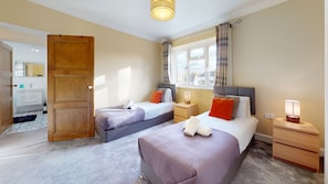 A hotel room with two beds, a wooden door, a window with curtains, a bedside table with a lamp, and a bathroom visible through an open door. - Immaculate 4-bed House in Romford (Romford)