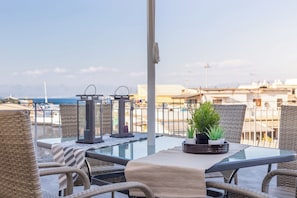 Mesa con cubierta de vidrio, sillas de mimbre, una planta en maceta y dos faroles en una terraza con vistas a edificios y un cuerpo de agua.