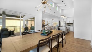 A spacious dining area with a wooden table and chairs, a grill outside, and a modern kitchen in the background. - Modern Lake Escape: Pool, Spa, Dock, Game Room, Mini Golf, Sleeps 29! (Lake Ozark)