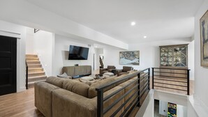 A spacious living area with a large sectional sofa, a flat-screen TV mounted on the wall, and a staircase leading to an upper level. - Modern Lake Escape: Pool, Spa, Dock, Game Room, Mini Golf, Sleeps 29! (Lake Ozark)