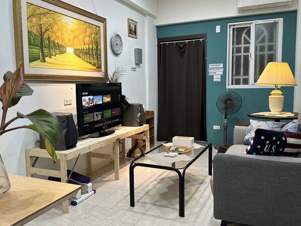 A living room with a TV, a sofa, a table, a fan, and a painting on the wall.