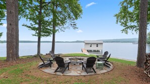 Un patio junto al lago con sillas Adirondack, una hoguera y vista a una casa al otro lado del agua.