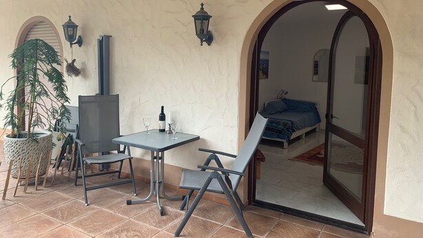 Eine Terrasse mit Tisch, Stühlen und einem Blumentopf, durch einen Bogen hindurch ist ein Bett zu sehen.