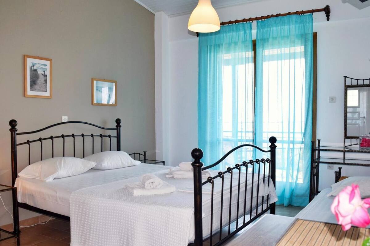 A double bed with white linens, a bedside table, a mirror, and a window with blue curtains.