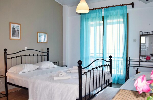 A double bed with white linens, a bedside table, a mirror, and a window with blue curtains.