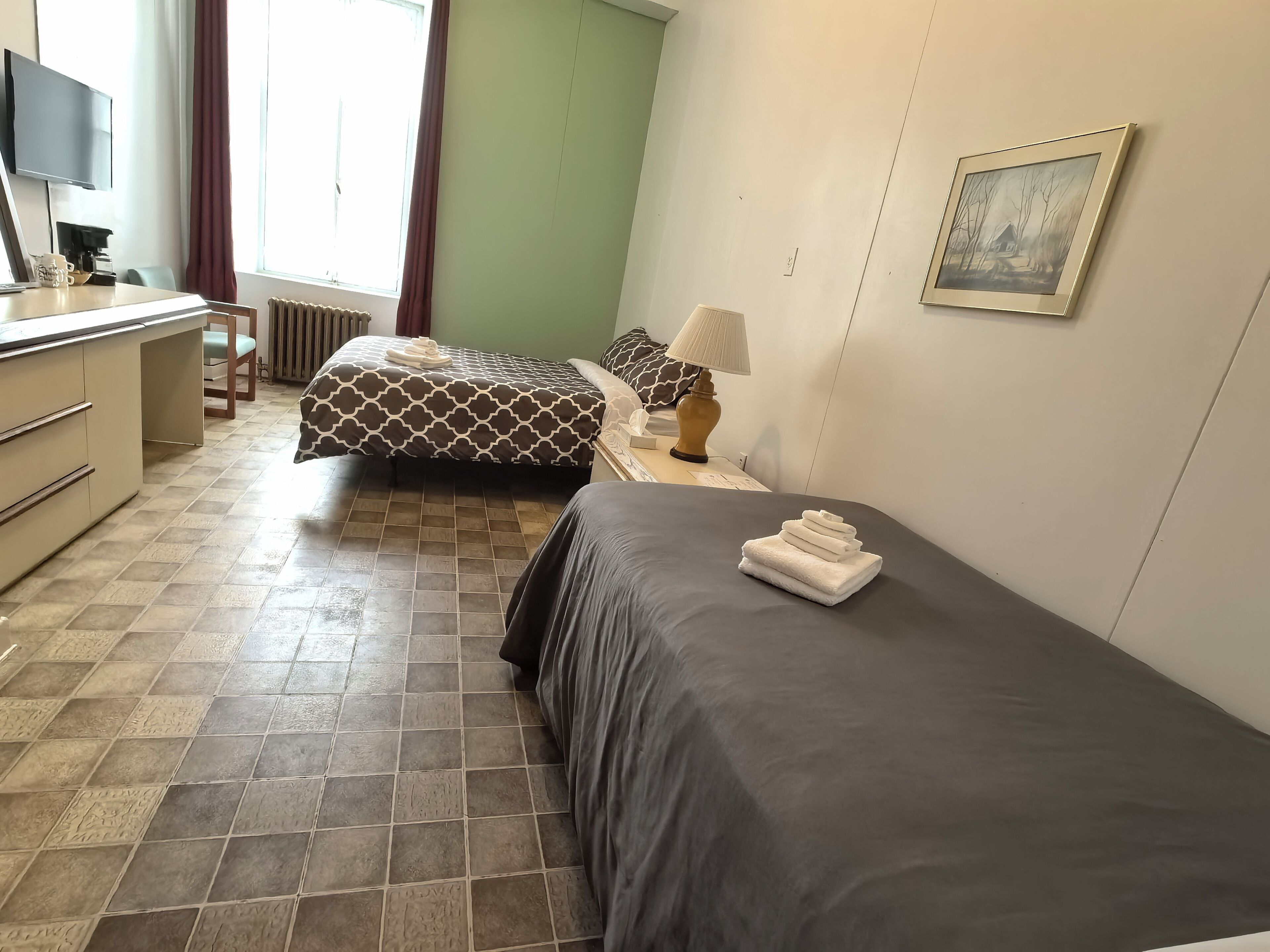 A hotel room with two beds, a desk, a chair, a radiator, a lamp, and a painting on the wall.