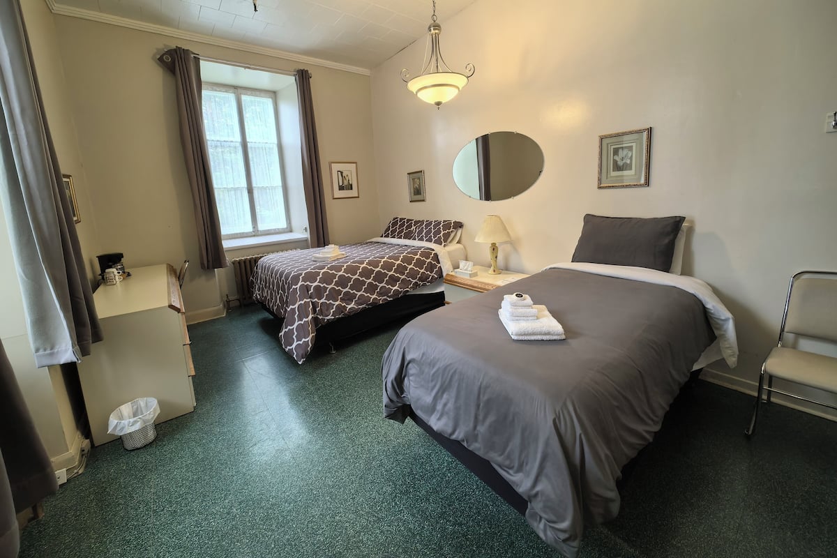 Habitación de hotel con dos camas, un escritorio, una silla, un ventanal con cortinas, un espejo y una lámpara en el techo.