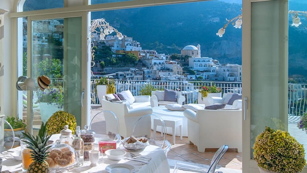Un coin repas avec une table dressée pour quatre personnes, donnant sur une ville côtière aux bâtiments blancs, avec une chaîne de montagnes en arrière-plan.