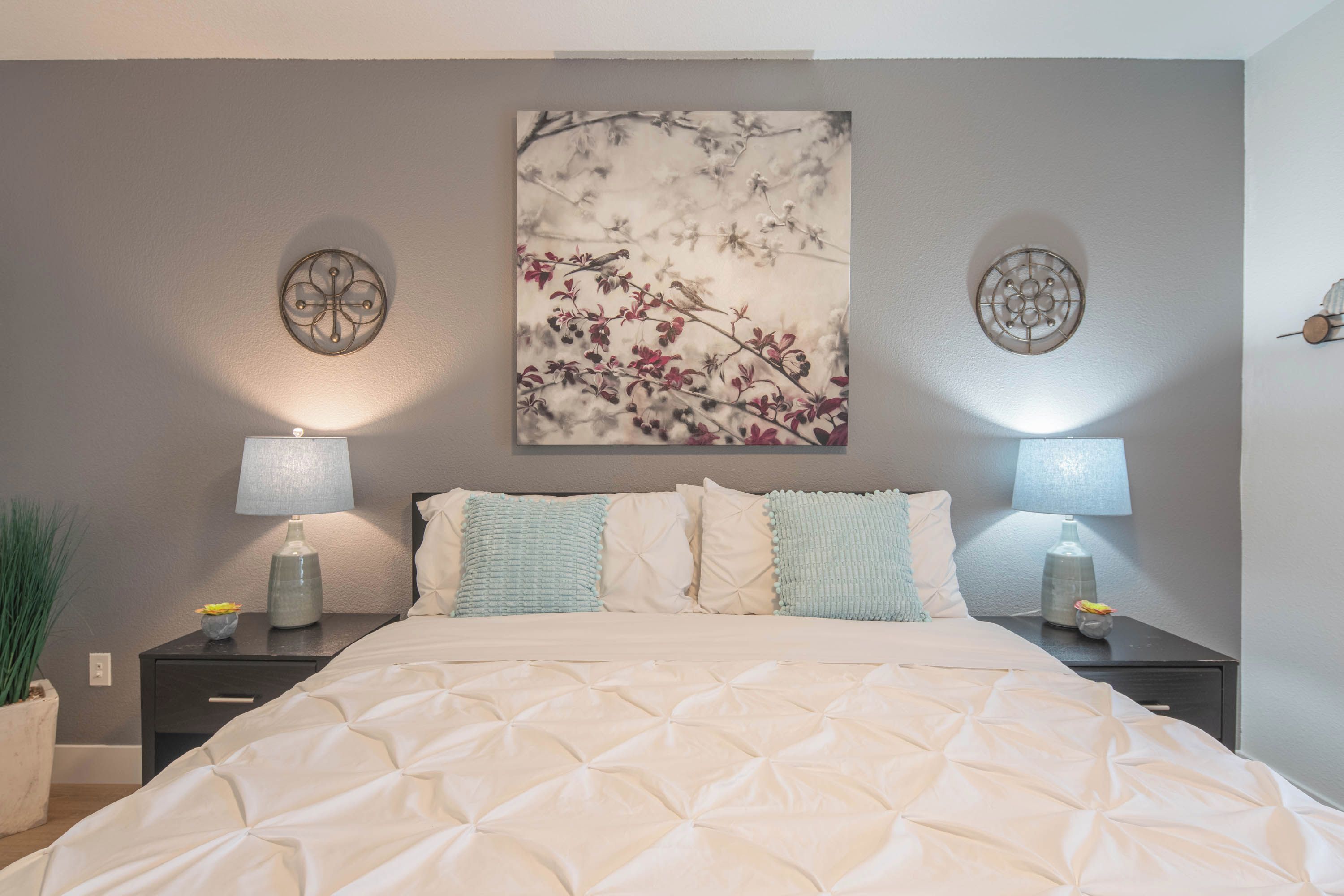 Una cama bien hecha con cubrecama acolchado, dos mesitas de noche con lámparas, una obra de arte en la pared que muestra una rama con bayas rojas, y una planta en maceta a la izquierda.