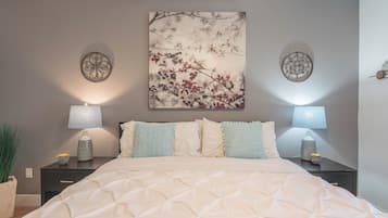 Una cama bien hecha con cubrecama acolchado, dos mesitas de noche con lámparas, una obra de arte en la pared que muestra una rama con bayas rojas, y una planta en maceta a la izquierda.