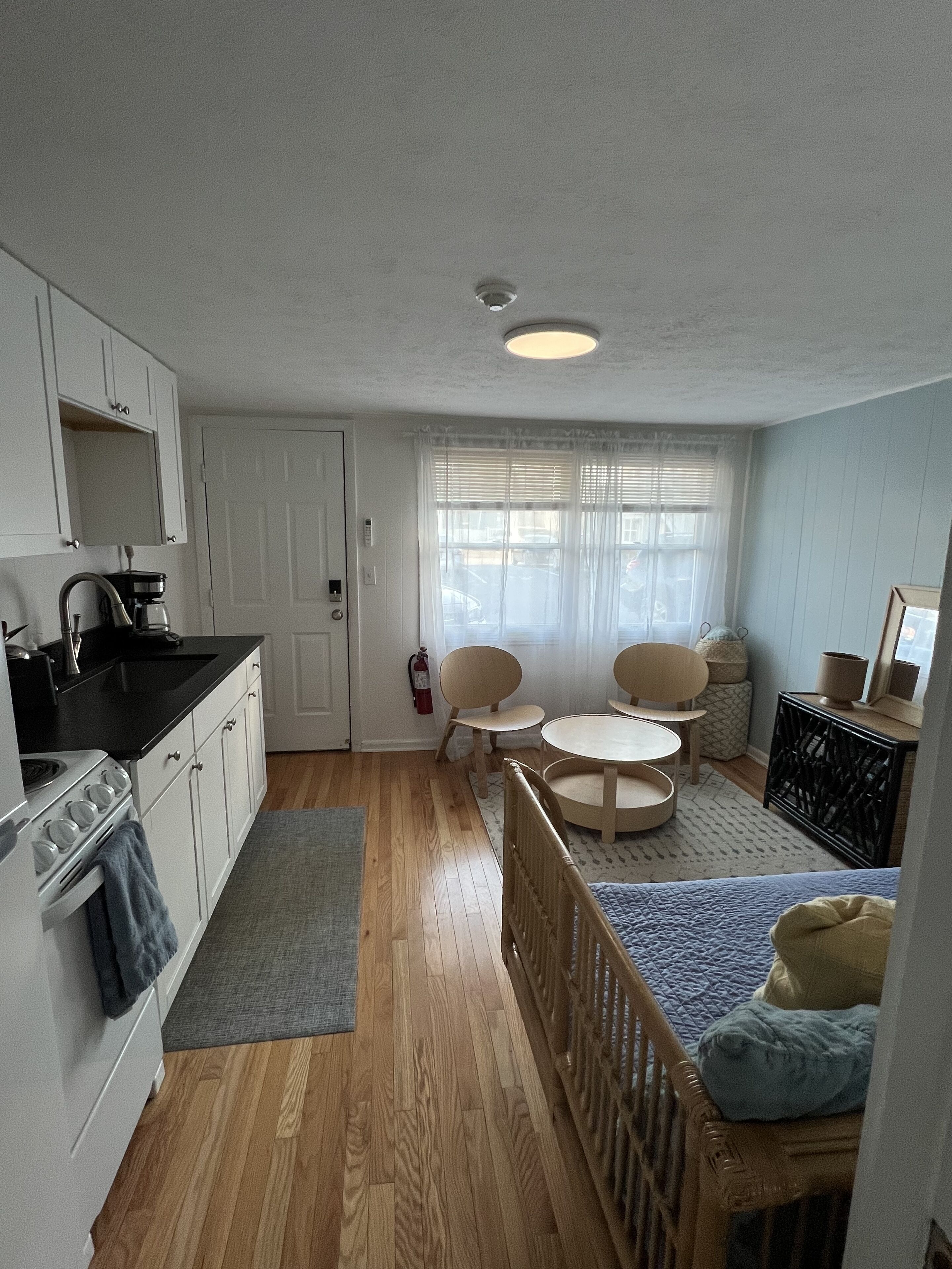 A compact kitchen with white cabinets, a stove, and a sink. There is a dining area with a small table and two chairs. A crib is visible in the corner.