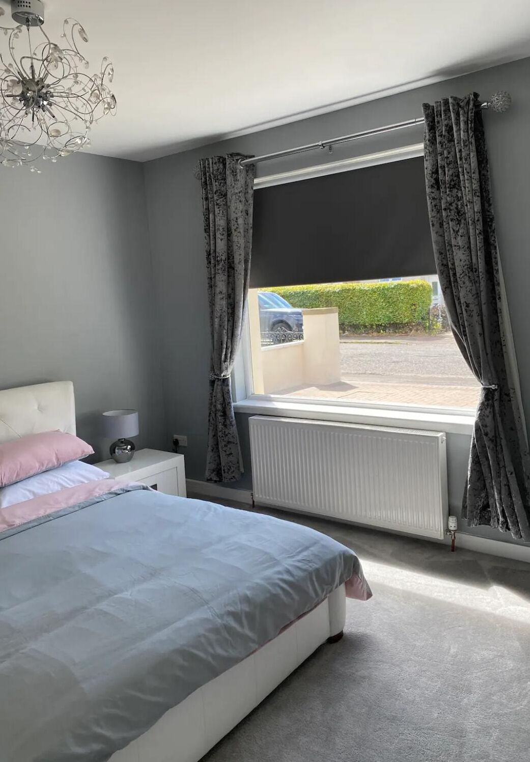 A bedroom with a bed, a nightstand, a lamp, a radiator, and a window with curtains.
