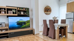 Ein modernes Wohnzimmer mit Flachbildfernseher, Holzboden, Essbereich mit Tisch und Stühlen sowie einer Küche mit Kühlschrank.