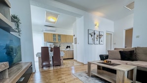 Ein modernes Wohnzimmer mit Couch, Fernseher auf Holzständer und Couchtisch. Im Hintergrund befindet sich eine Küche mit Holzschränken und ein Essbereich mit zwei Stühlen.