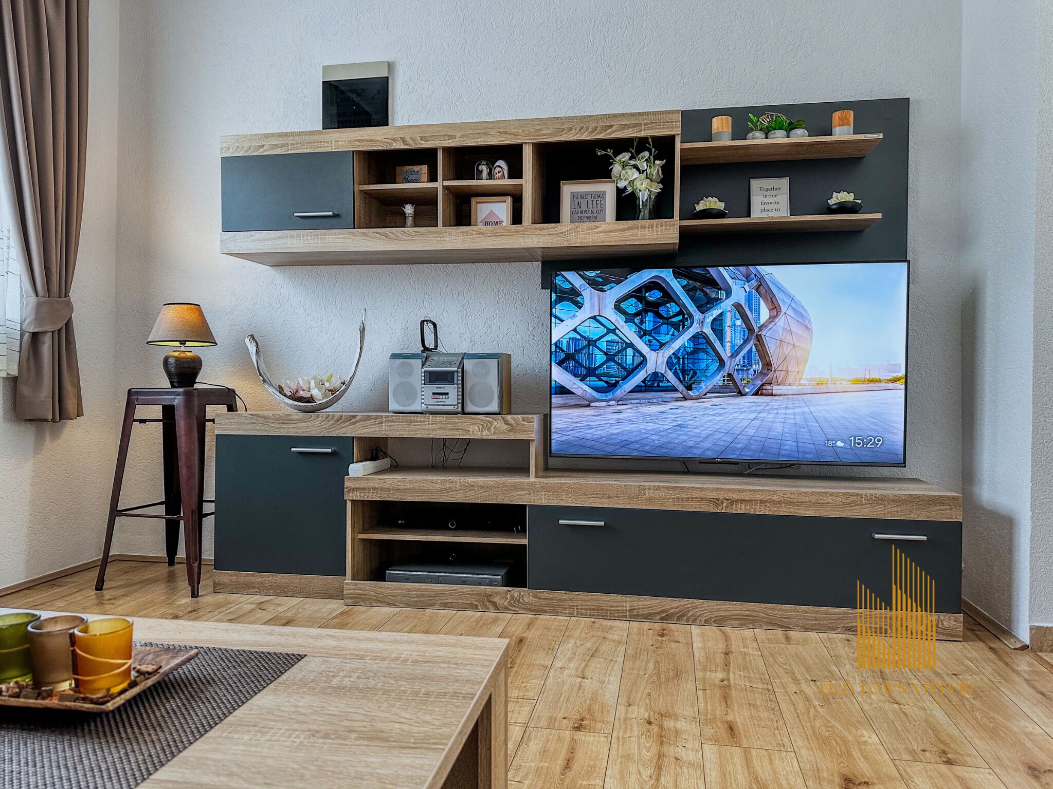 Moderni olohuone, jossa on suuri litteänäyttöinen televisio, puuhyllyt, pieni pöytä lampun kanssa ja koristeellinen kulho.