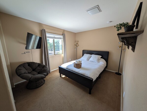 A bedroom with a bed, a chair, a TV, a window, and a plant.