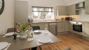 Une cuisine moderne avec un coin repas, comprenant une table en bois dressée pour deux personnes, un four encastré et une fenêtre équipée de stores.