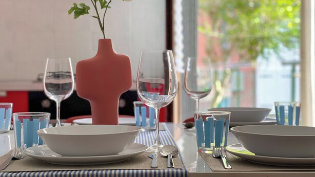 A dining table set with white plates, blue-striped placemats, and clear glassware. A pink vase with white flowers is placed on the table. - Apartment for 9 people - 4 min walk from Mestre Station (Venice) (Venezia)