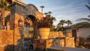 Un muro de piedra con una reja de hierro forjado, una jardinera grande con flores rojas y una palmera al fondo.