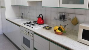 Une cuisine avec des meubles blancs, une bouilloire rouge, une cuisinière à gaz, un évier et un panier de fruits posé sur le plan de travail.