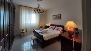 Une chambre d’hôtel avec un lit, un bureau, une chaise, une lampe, un tableau au mur et une fenêtre avec des rideaux.
