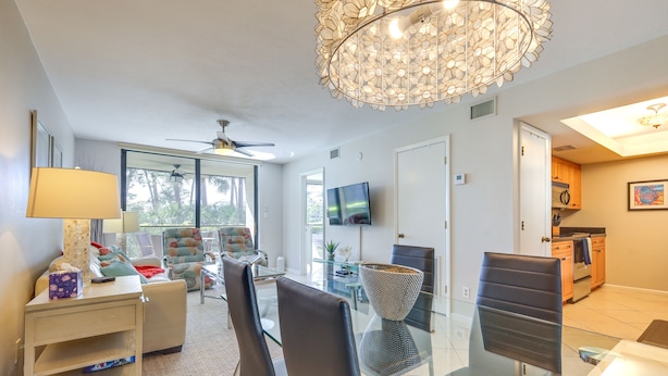 Ein modernes Wohnzimmer mit einem Glastisch, gemütlicher Sitzgelegenheit, einer Chandelierleuchtre und einem an der Wand befestigten Fernseher.