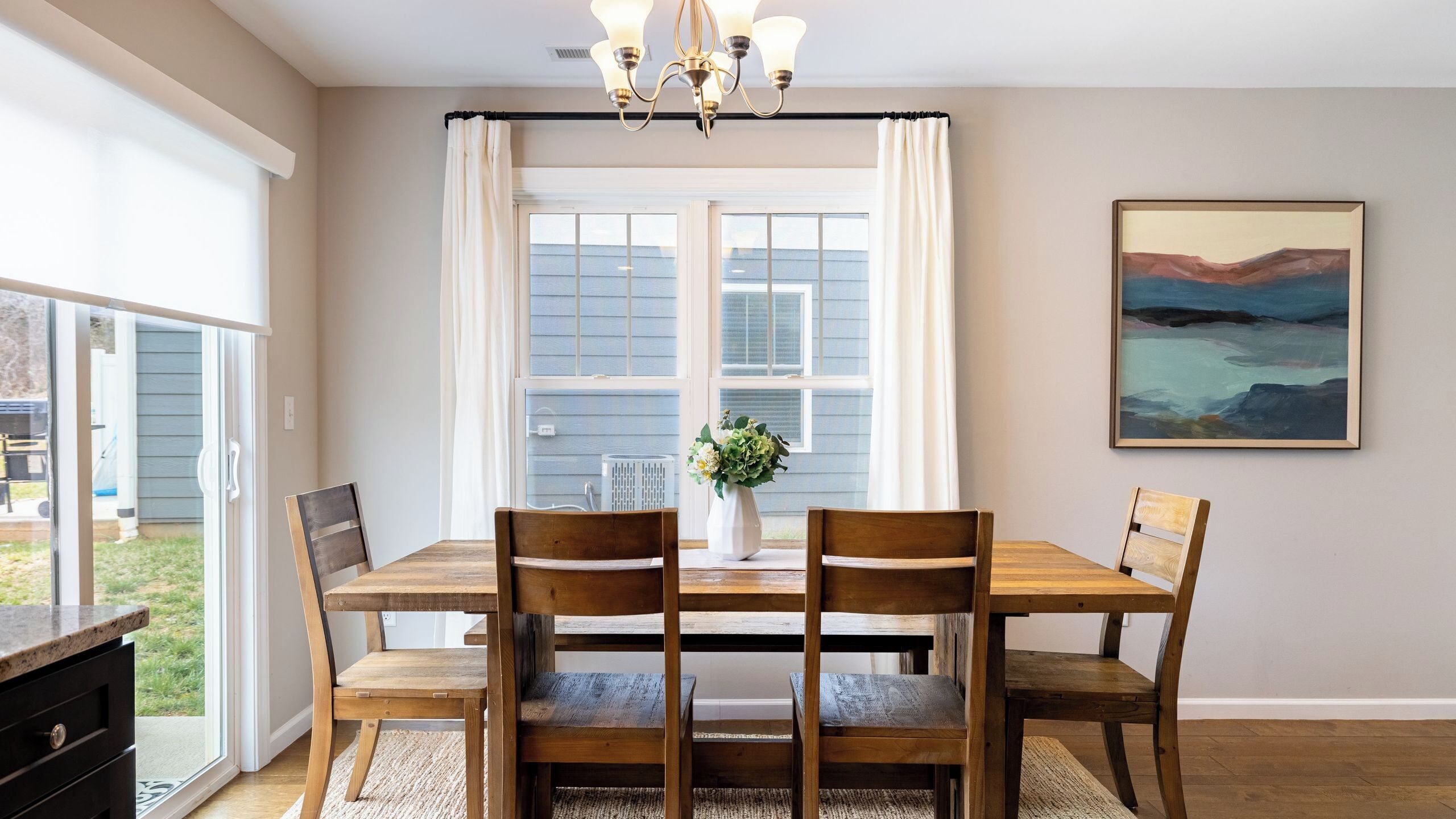A dining area with a wooden table, four chairs, a vase with flowers, and a painting on the wall.