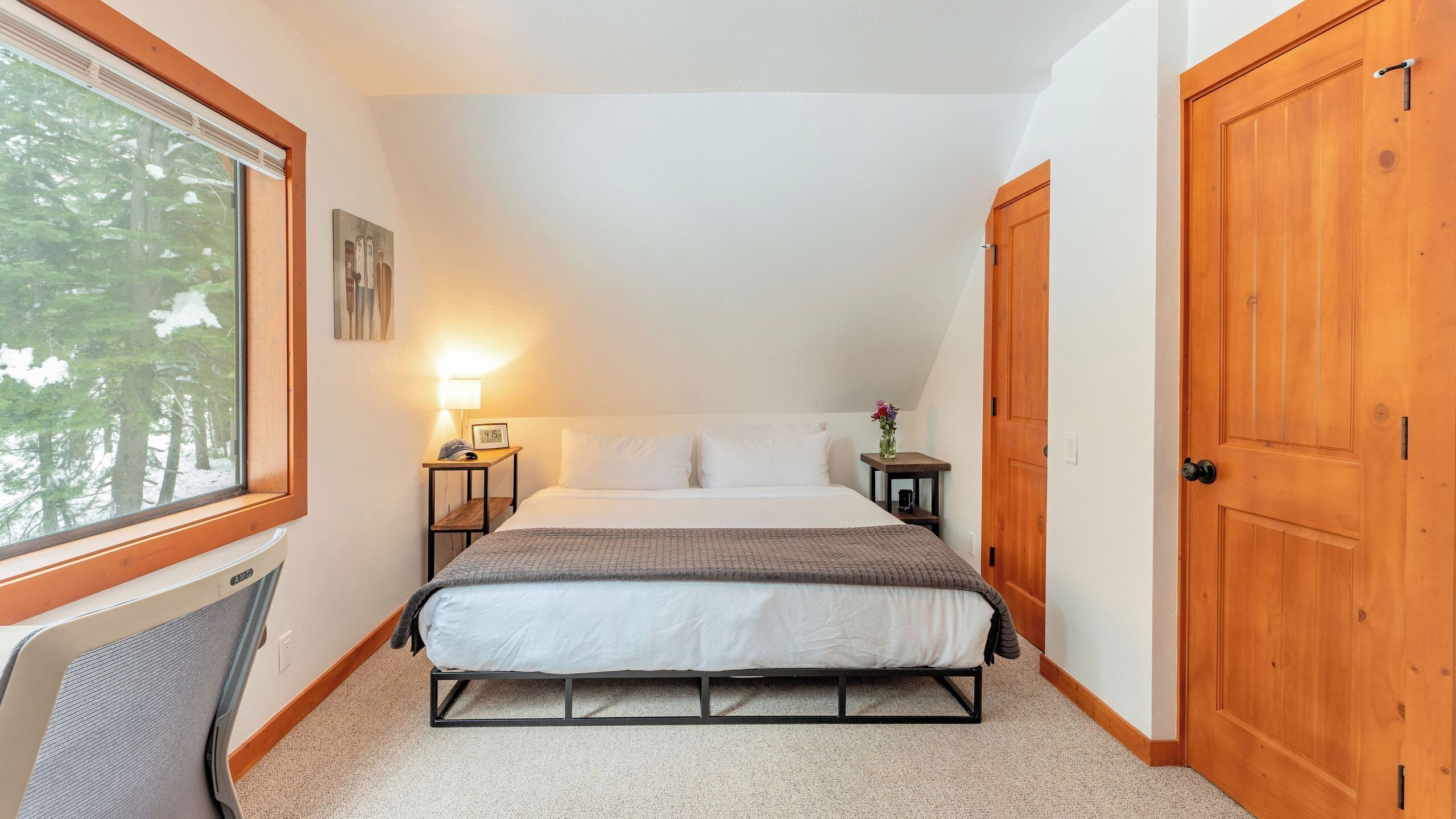 Un dormitorio con cama, una mesita de noche con lámpara, una ventana con vistas a árboles y una puerta de madera.